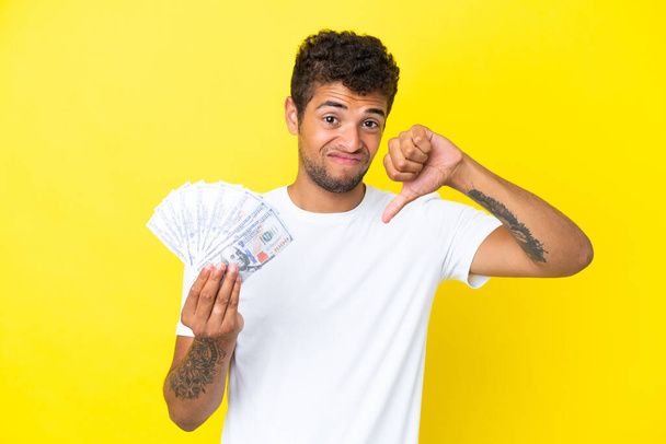 Mladý brazilský muž brát spoustu peněz izolovaných na žlutém pozadí ukazující palec dolů s negativním výrazem - Fotografie, Obrázek