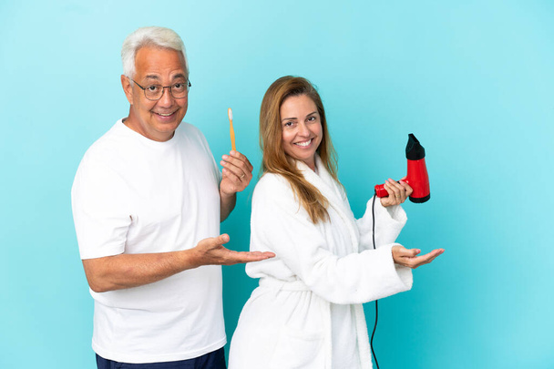 Пара средних лет, держащая сушилку и зубную щетку изолированы на синем фоне протягивая руки в сторону для приглашения приехать - Фото, изображение