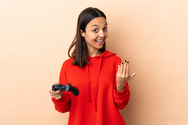 Молода брюнетка змішаної раси жінка грає з контролером відеоігор над ізольованим фоном, запрошуючи прийти
 - Фото, зображення