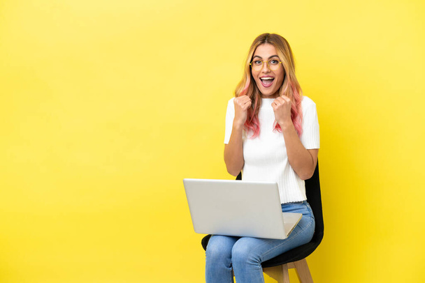 Jovem mulher sentada em uma cadeira com laptop sobre fundo amarelo isolado celebrando uma vitória na posição de vencedora - Foto, Imagem