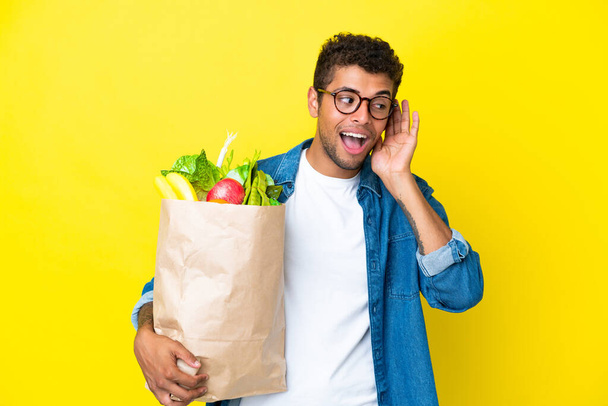 Junger brasilianischer Mann mit einer Einkaufstasche in der Hand, isoliert auf gelbem Hintergrund, hört etwas, indem er die Hand ans Ohr legt - Foto, Bild