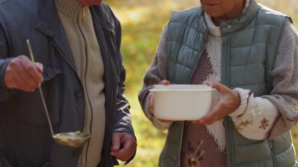 Bienestar social. Evento de caridad. Hombre vertiendo sopa caliente en un recipiente de plástico en las manos de una anciana - Metraje, vídeo