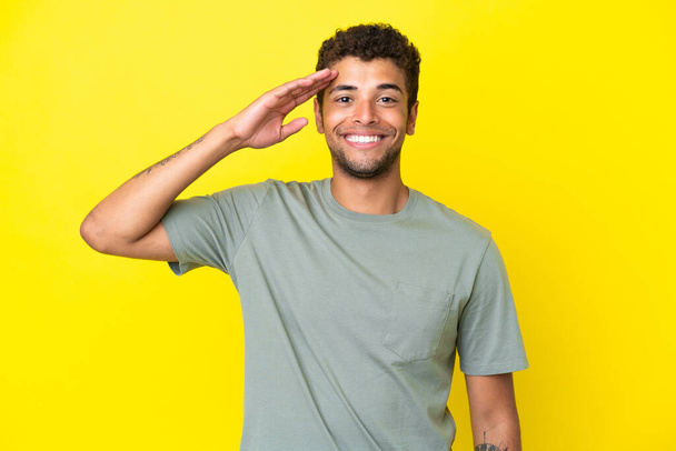 Young handsome Brazilian man isolated on yellow background saluting with hand with happy expression - Fotoğraf, Görsel