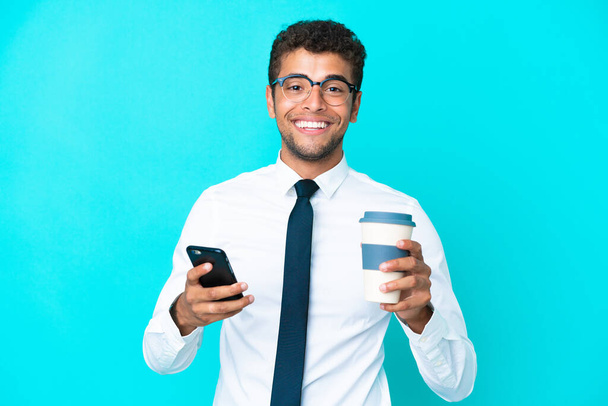 Joven empresario brasileño aislado sobre fondo azul sosteniendo café para llevar y un móvil - Foto, Imagen