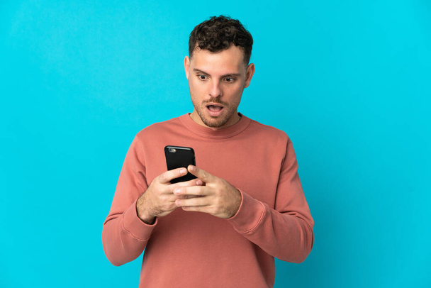 Young caucasian handsome man isolated on blue background looking at the camera while using the mobile with surprised expression - Fotografie, Obrázek