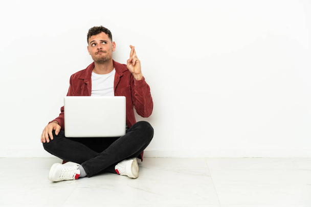 Jonge knappe blanke man sit-in op de vloer met laptop met vingers kruisen en wensen de beste - Foto, afbeelding