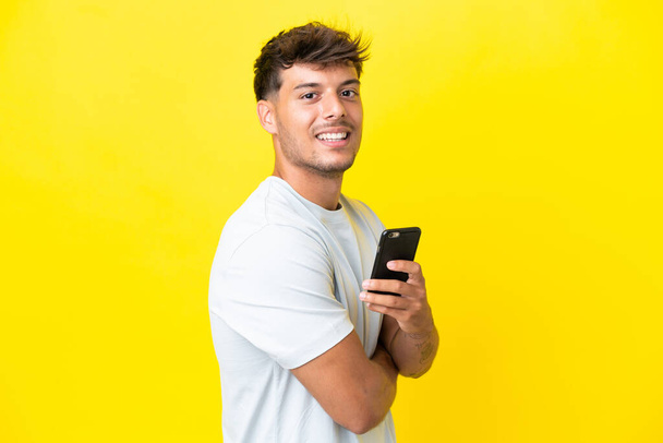 Joven hombre guapo caucásico aislado sobre fondo amarillo sosteniendo un teléfono móvil y con los brazos cruzados - Foto, Imagen