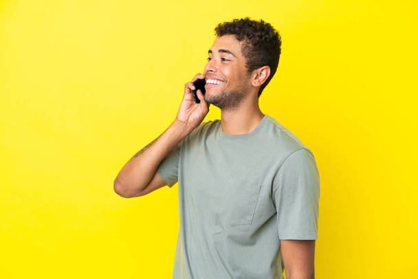 Jovem brasileiro bonito isolado em fundo amarelo mantendo uma conversa com o celular - Foto, Imagem