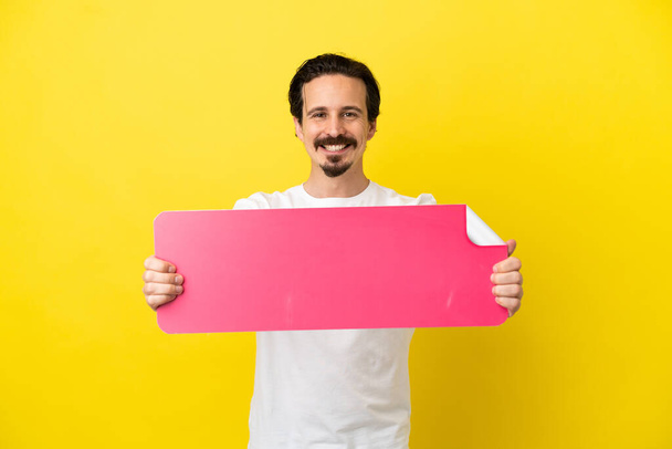Young caucasian man isolated on yellow background holding an empty placard - 写真・画像