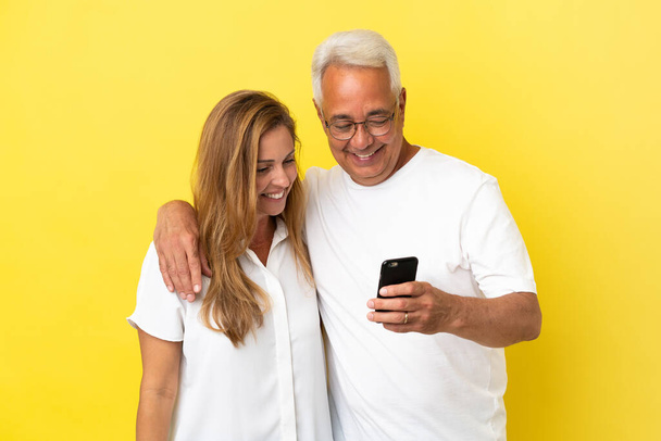 Pareja de mediana edad aislada sobre fondo amarillo enviando un mensaje o correo electrónico con el móvil - Foto, imagen