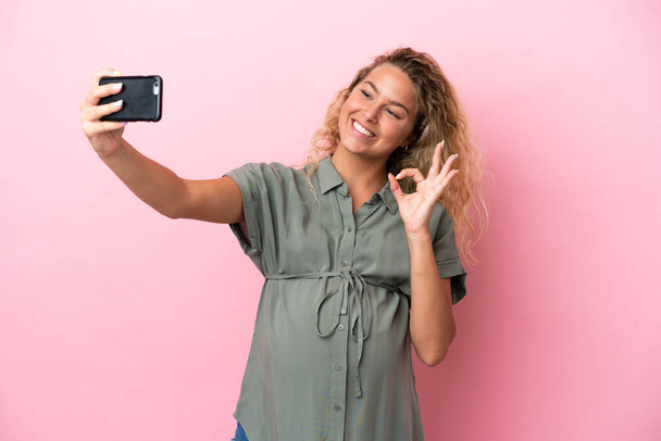 Menina com cabelo encaracolado isolado no fundo rosa grávida e fazendo uma selfie - Foto, Imagem