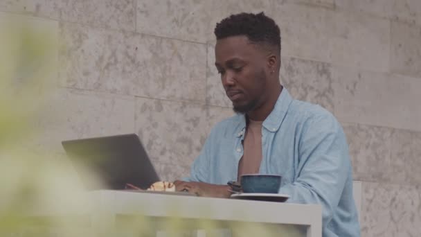 屋外のレストランのテーブルに座っている若い黒人男性の傾き,朝食にポータブルコンピュータを使用して,コーヒーを飲む - 映像、動画