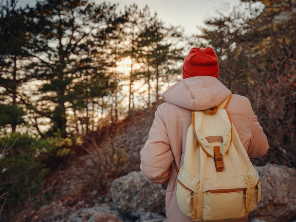 Człowiek z plecakiem i czerwonym kapeluszem wędrujący w górach zachodzącego słońca stoi na skalistym szlaku turystycznym lub ścieżce. Zimna pogoda. Zimowa wędrówka. Wibracje i przygody na świeżym powietrzu - Zdjęcie, obraz