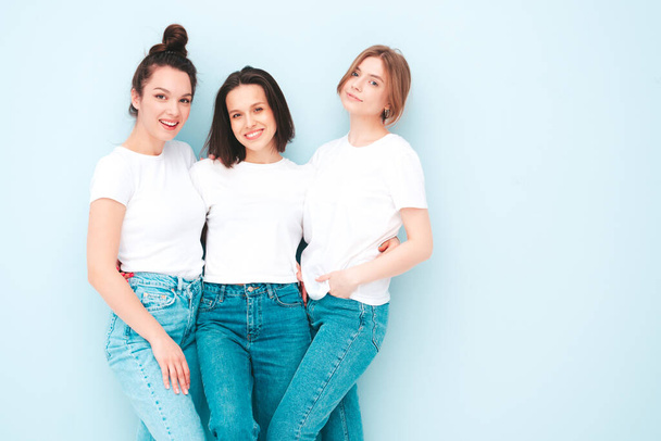 Three young beautiful smiling hipster female in trendy same summer white t-shirt and jeans clothes. Sexy carefree women posing near light blue wall in studio. Cheerful and positive models having fun - Φωτογραφία, εικόνα