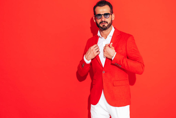 Portrait of handsome confident stylish hipster lambersexual model. Sexy modern man dressed in elegant suit. Fashion male posing in studio near red wall in sunglasses - Zdjęcie, obraz