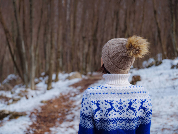 Beautiful woman standing among trees in winter forest. Wearing hat and blue sweater. Caucasian Asian female model outside in first snow - Photo, Image