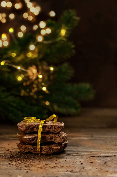 Pieces of chocolate on a wooden table with selective blurred focus on the background of fir branches with lights. Close-up of pieces of chocolate with copy space. - Photo, Image