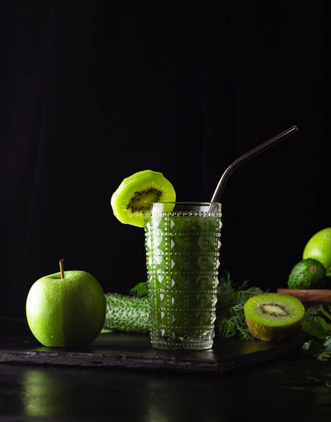 Green smoothie in a glass glass on a black background. Kiwi, apples,cucumbers and greens. Cooking healthy food. Zero weist, a metal tube. - Foto, afbeelding