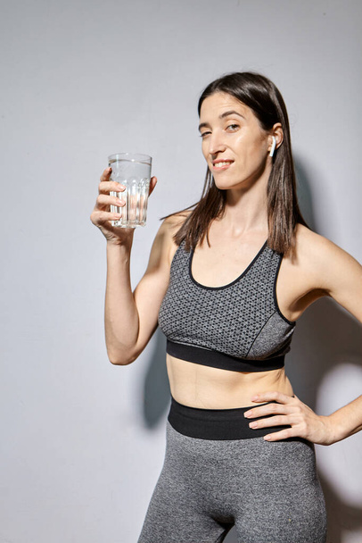 Eine Frau in Sportkleidung und mit drahtlosen Kopfhörern trinkt Wasser. das Konzept eines gesunden Lebensstils und sportlicher Aktivitäten - Foto, Bild