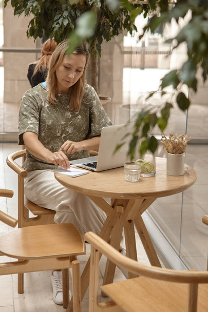 Egy fiatal nő ül egy kávézóban, és egy laptopon dolgozik. A szabadúszó és távoli munkavégzés vagy képzés fogalma. Egy nő jegyzetel egy jegyzetfüzetben, és megnézi a laptop képernyőjét. - Fotó, kép