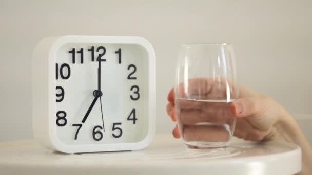 White alarm clock and glass of water on table in bedroom. Hand holding glass, close up view. Wake up early in the morning and drink water. Good habits for healthy way of life, dehydration concept - Footage, Video