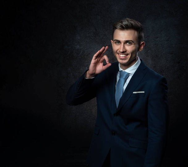 A young male steward, in a suit, smiles, makes a hand gesture everything is ok. Against a dark background. - Фото, зображення