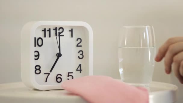 White alarm clock and glass of water on table in bedroom. Hand holding glass, close up view. Wake up early in the morning and drink water. Good habits for healthy way of life, dehydration concept - Footage, Video