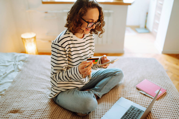 Online shopping t home. A young woman holds a credit card and uses a laptop. Black friday, sale, consumerist, lifestyle concept. - Фото, изображение