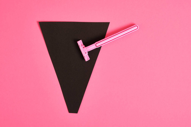 a pink plastic disposable razor and a black triangle made of paper on a pink background, an association on the theme of feminism and shaving hair from a female body - Foto, immagini
