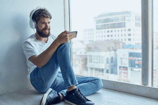 vousatý muž sedí u okna ve sluchátkách a poslouchá hudbu na sluchátkách komunikace - Fotografie, Obrázek