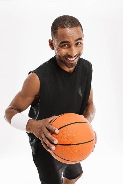 Jonge zwarte sportman glimlachen op camera terwijl poseren met basketbal geïsoleerd over witte achtergrond - Foto, afbeelding