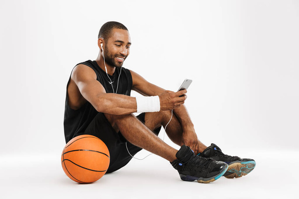Feliz jugador de baloncesto africano joven sentado escuchando música con auriculares y teléfono móvil aislado sobre un fondo blanco - Foto, Imagen