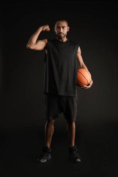 Young sportsman showing his bicep while posing with basketball isolated over black background - Photo, Image