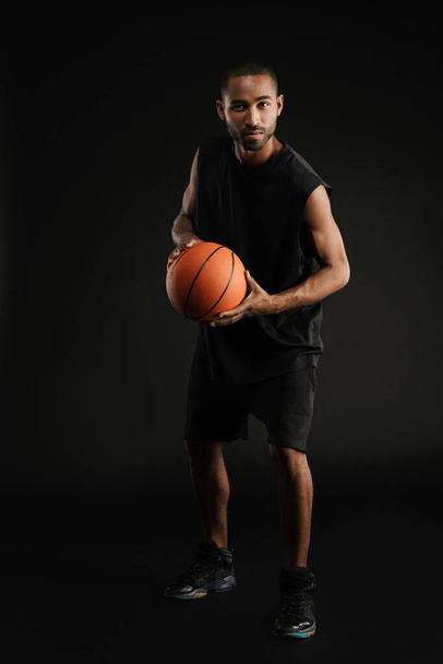 Young sportsman looking at camera while posing with basketball isolated over black background - 写真・画像