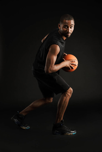 Young bristle sportsman playing basketball while working out isolated over black background - Zdjęcie, obraz