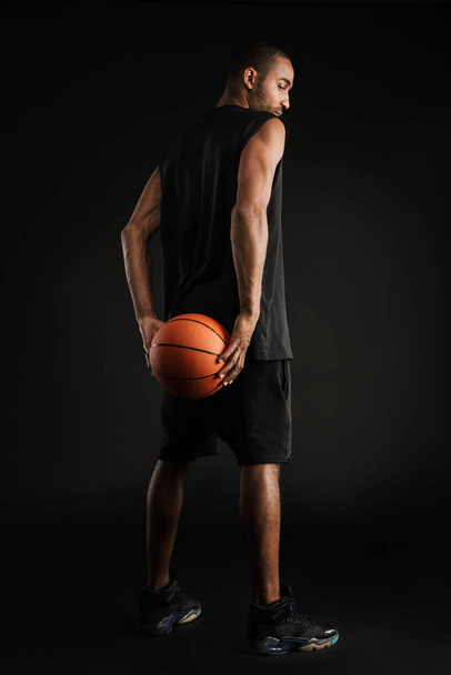 Young sportsman looking downward while posing with basketball isolated over black background - Foto, imagen