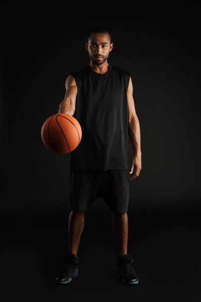 Young sportsman looking and reaching basketball at camera isolated over black background - Fotografie, Obrázek