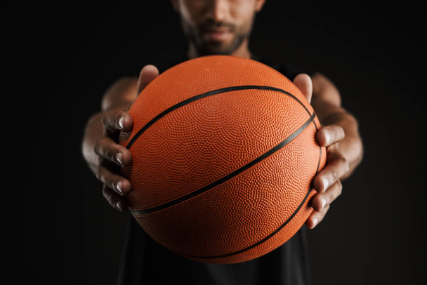Young sportsman looking and reaching basketball at camera isolated over black background - 写真・画像