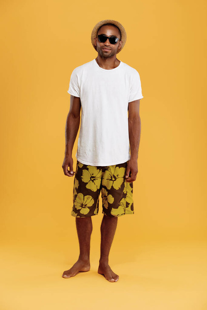 Přední pohled na pohledného mladého černocha. Chlápek nosí tričko, plavky, slamák a brýle. Koncept letní dovolené na pláži. Izolováno na žlutém pozadí. Natáčení. Kopírovat prostor - Fotografie, Obrázek