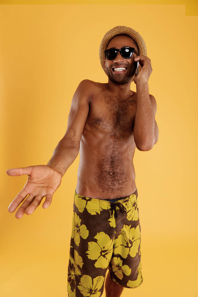 Young pleased handsome black man talking on mobile phone. Guy wear swimming trunks, straw hat and glasses. Concept of summer beach vacation. Isolated on yellow background. Studio shoot - Foto, immagini