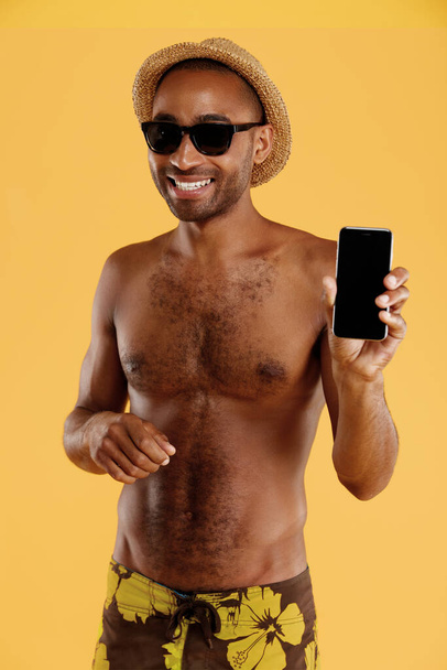 Young smiling handsome black man showing mobile phone. Guy wear swimming trunks, straw hat and glasses. Concept of summer beach vacation. Isolated on yellow background. Studio shoot - Fotografie, Obrázek