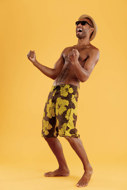 Young successful good-looking black man celebrating. Guy wear swimming trunks, straw hat and glasses. Concept of summer beach vacation. Isolated on yellow background. Studio shoot - Fotoğraf, Görsel