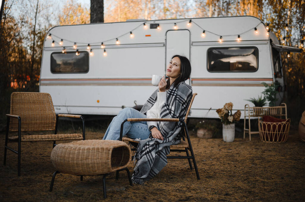 Beautiful caucasian woman is sitting wrapped in a plaid and drinking a warming drink outdoors. Traveling in a motor home in autumn. - Foto, immagini