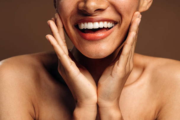 Cropped view of the overjoyed multiracial brunette girl with perfect skin and nude makeup smiling toothy while posing over brown studio background - Foto, imagen