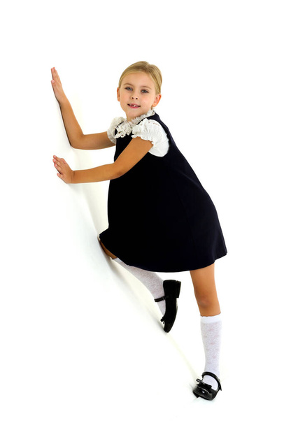 A cheerful blonde in a white blouse and a blue dress puts her hands on the wall. Lovely seven year old child standing against white background - 写真・画像