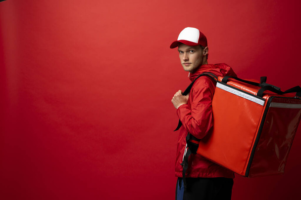 Portrait of a young courier, delivery man in red uniform with a thermal backpack isolated on a red background. Fast home delivery. Online order. Courier delivers groceries home. - Foto, Imagem