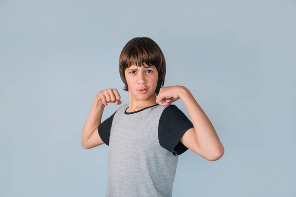 Portret van tiener jongen Flexen zijn biceps met brutaal plezier blik op zijn gezicht blauwe achtergrond met kopieerruimte - Foto, afbeelding