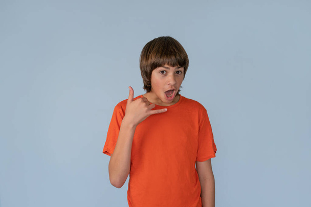 Fun Portret van tiener jongen doet een Call Me Gesture met Brutaal Verrast blik op zijn gezicht blauwe achtergrond met Copy Space - Foto, afbeelding
