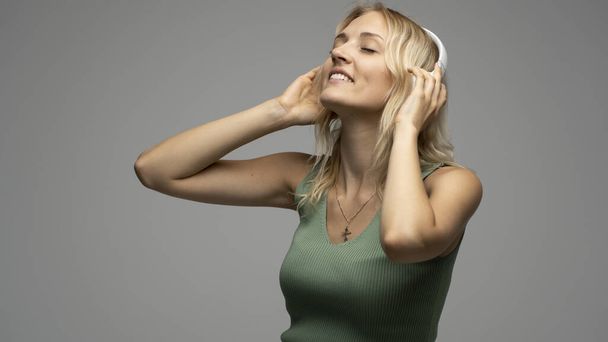 Beautiful attractive young blond woman wearing green t-shirt and glasses in white headphones listening music and smiling on grey background in studio. Relaxing and enjoying. Lifestyle. - Valokuva, kuva