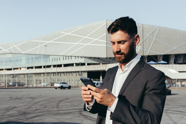 Portrait of confident businessman in a classic suit having video call with business partner using mobile phone standing near modern office building and white car. Technologies concept. - Zdjęcie, obraz